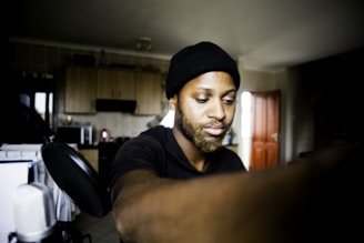 A person wearing a black beanie is seated in a room with an open wooden door in the background. The room contains kitchen elements such as a microwave and cabinets. A large microphone is visible on the left, suggesting a podcast or music recording setting. The lighting is soft, casting subtle shadows.