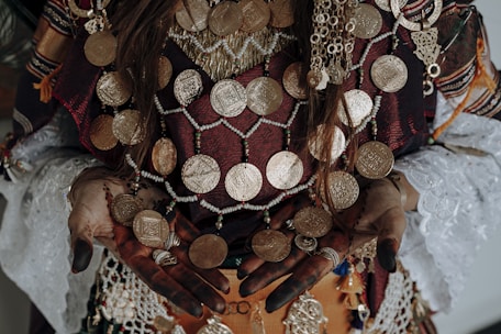 Ornate jewelry with large gold coins adorns a person wearing traditional attire. The intricate design includes beads and various patterns on fabric, with henna-decorated hands showing rings and accessories. The textiles feature rich textures and colors.