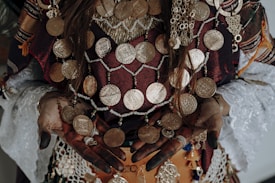 Ornate jewelry with large gold coins adorns a person wearing traditional attire. The intricate design includes beads and various patterns on fabric, with henna-decorated hands showing rings and accessories. The textiles feature rich textures and colors.