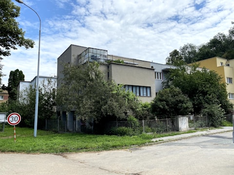 A modern, minimalist building is surrounded by lush greenery on a partly cloudy day. The structure features flat roofs, clean lines, and some glass elements, typical of contemporary architecture. The foreground includes a well-maintained lawn, a street lamp, and a speed limit sign indicating 30 km/h.