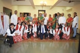 A group of adults and children posed together in traditional attire. The children are dressed in coordinated red and white outfits with hats, while the adults are in colorful traditional sarees and other attire. The setting appears to be indoors with a chandelier overhead and clothing racks to one side.
