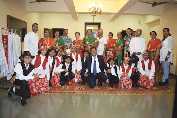 A group of adults and children posed together in traditional attire. The children are dressed in coordinated red and white outfits with hats, while the adults are in colorful traditional sarees and other attire. The setting appears to be indoors with a chandelier overhead and clothing racks to one side.