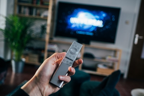 A hand holding a remote control points towards a large TV screen with the Universal logo displayed. The room includes a wooden shelf with books and a plant, and appears cozy and lived-in.