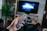 A hand holding a remote control points towards a large TV screen with the Universal logo displayed. The room includes a wooden shelf with books and a plant, and appears cozy and lived-in.