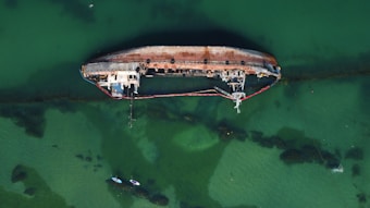 An aerial view of a large, rusted shipwreck partially submerged in greenish water. The ship lies on its side and is surrounded by a thin red containment boom. The water around the ship is shallow, with visible shadows and darker patches indicating submerged structures or debris. Two small boats or kayaks are near the bottom of the image, providing a sense of scale.