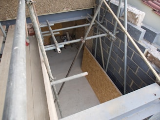Scaffolding is set up around a construction area with partially finished walls and exposed building materials. The scene includes metal poles, wooden boards, and a concrete floor. Some construction debris is scattered on the ground.