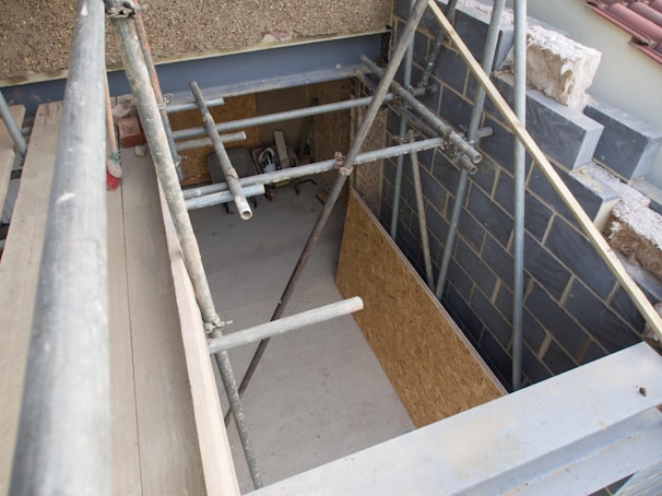Scaffolding is set up around a construction area with partially finished walls and exposed building materials. The scene includes metal poles, wooden boards, and a concrete floor. Some construction debris is scattered on the ground.