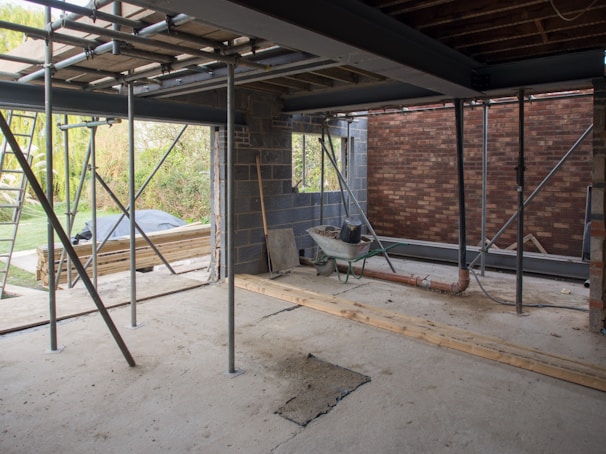 A construction site in progress with exposed steel beams, scaffolding, and a partially built brick wall. A wheelbarrow and various construction materials are scattered on the unfinished concrete floor. Wooden planks and a ladder are seen, with greenery visible in the background through an opening.