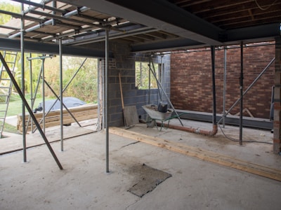 A construction site in progress with exposed steel beams, scaffolding, and a partially built brick wall. A wheelbarrow and various construction materials are scattered on the unfinished concrete floor. Wooden planks and a ladder are seen, with greenery visible in the background through an opening.