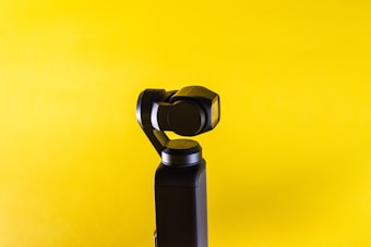 A black handheld stabilizer or gimbal with a camera is positioned against a bright yellow background. The device features a sleek, modern design with smooth, rounded edges.