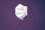 A single white disposable face mask with ear loops is suspended against a solid purple background. The mask appears slightly crumpled and is positioned at an angle, with the ear loops hanging loosely.