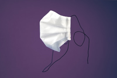 A single white disposable face mask with ear loops is suspended against a solid purple background. The mask appears slightly crumpled and is positioned at an angle, with the ear loops hanging loosely.