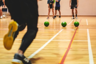 A group of people in a gym setting appear engaged in a friendly dodgeball game. Three green balls are lined up on the wooden floor as a player wearing black pants and athletic shoes runs towards them. Other players are visible in the background, casually observing or preparing to participate.