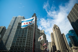 The image depicts an urban cityscape with tall buildings against a bright blue sky. Multiple flags are seen, including one prominently displaying the distinctive design with stars and stripes. The skyscrapers vary in architectural style, adding to the city&rsquo;s skyline.