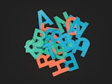A pile of colorful, cut-out cardboard letters in shades of red, blue, and green against a dark background, arranged in a random fashion with overlapping and tilted angles.