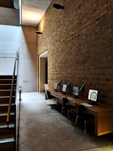 A modern interior space featuring a tall brick wall and a polished concrete floor. Alongside the wall, there is a wooden desk with four computer workstations, each equipped with a sleek black chair. The area is illuminated by soft, ambient lighting, which casts shadows on the wall. A staircase with metal railings is visible on the left, leading up to another level.
