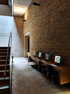 A modern interior space featuring a tall brick wall and a polished concrete floor. Alongside the wall, there is a wooden desk with four computer workstations, each equipped with a sleek black chair. The area is illuminated by soft, ambient lighting, which casts shadows on the wall. A staircase with metal railings is visible on the left, leading up to another level.