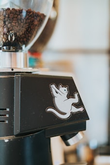 A coffee grinder featuring a transparent hopper filled with roasted coffee beans. The grinder is adorned with a playful sticker of a cartoon cat in a striking pose.