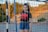 A girl stands in front of a sports goal on a field with a scenic background of trees and a hill. She wears a black t-shirt with red text and denim shorts, with her hair tied back in a ponytail.
