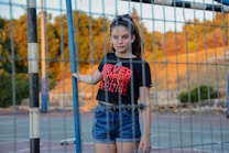A girl stands in front of a sports goal on a field with a scenic background of trees and a hill. She wears a black t-shirt with red text and denim shorts, with her hair tied back in a ponytail.