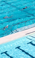 Several swimmers are in a large outdoor swimming pool with clear blue water. The pool has lanes marked by ropes, and the sunlight creates patterns on the water surface, giving a sense of movement and fluidity.