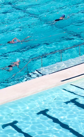 Several swimmers are in a large outdoor swimming pool with clear blue water. The pool has lanes marked by ropes, and the sunlight creates patterns on the water surface, giving a sense of movement and fluidity.