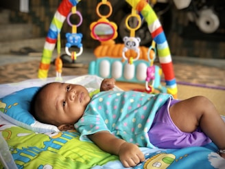 A baby lies on a colorful play mat, wearing a polka dot shirt and purple shorts. Above the baby is an activity gym featuring hanging toys in various shapes and colors including rings and plush animals. This setup is on a patterned surface, and the background includes steps and part of a bike or vehicle.