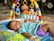 A baby lies on a colorful play mat, wearing a polka dot shirt and purple shorts. Above the baby is an activity gym featuring hanging toys in various shapes and colors including rings and plush animals. This setup is on a patterned surface, and the background includes steps and part of a bike or vehicle.