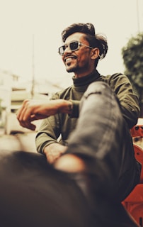 A person wearing sunglasses and casual clothing is smiling confidently. The setting appears urban with a blurred background that includes buildings and greenery. The angle is slightly low, emphasizing the relaxed and cheerful demeanor.