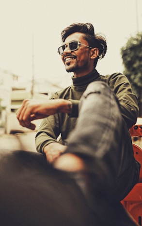 A person wearing sunglasses and casual clothing is smiling confidently. The setting appears urban with a blurred background that includes buildings and greenery. The angle is slightly low, emphasizing the relaxed and cheerful demeanor.