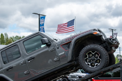 A gray Jeep Rubicon is positioned on an inclined ramp, demonstrating its off-road capabilities. In the background, a large American flag waves against a cloudy sky. There are banners advertising a lifetime limited warranty, and tree foliage is visible, suggesting an outdoor setting.