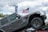 A gray Jeep Rubicon is positioned on an inclined ramp, demonstrating its off-road capabilities. In the background, a large American flag waves against a cloudy sky. There are banners advertising a lifetime limited warranty, and tree foliage is visible, suggesting an outdoor setting.