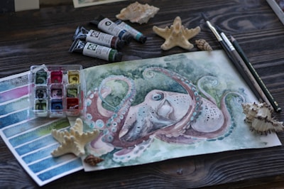 A watercolor painting of an octopus surrounded by art supplies including paint tubes, a container of watercolor paints, brushes, pencils, and seashells on a wooden surface. The painting itself features an intricate design with green, blue, and pink hues.