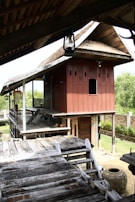 A traditional wooden house on stilts with a sloping roof and intricate carvings on the walls. The house features a raised wooden platform with steps leading up to it. The surrounding area includes lush greenery and a brick wall. An earthen pot is visible near the wooden structure.