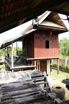 A traditional wooden house on stilts with a sloping roof and intricate carvings on the walls. The house features a raised wooden platform with steps leading up to it. The surrounding area includes lush greenery and a brick wall. An earthen pot is visible near the wooden structure.