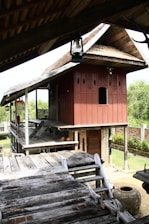 A traditional wooden house on stilts with a sloping roof and intricate carvings on the walls. The house features a raised wooden platform with steps leading up to it. The surrounding area includes lush greenery and a brick wall. An earthen pot is visible near the wooden structure.
