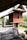 A traditional wooden house on stilts with a sloping roof and intricate carvings on the walls. The house features a raised wooden platform with steps leading up to it. The surrounding area includes lush greenery and a brick wall. An earthen pot is visible near the wooden structure.
