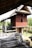 A traditional wooden house on stilts with a sloping roof and intricate carvings on the walls. The house features a raised wooden platform with steps leading up to it. The surrounding area includes lush greenery and a brick wall. An earthen pot is visible near the wooden structure.
