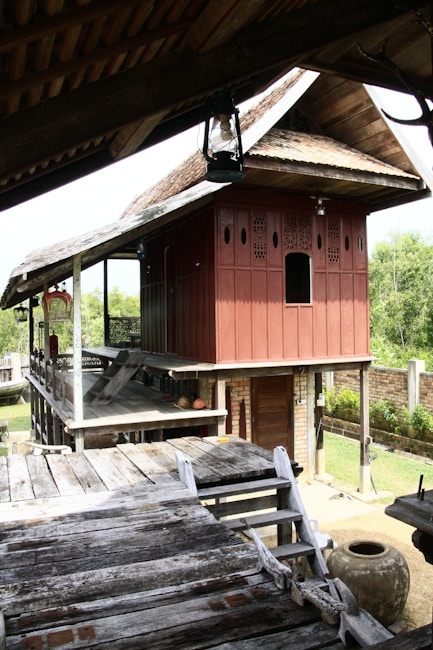 A traditional wooden house on stilts with a sloping roof and intricate carvings on the walls. The house features a raised wooden platform with steps leading up to it. The surrounding area includes lush greenery and a brick wall. An earthen pot is visible near the wooden structure.