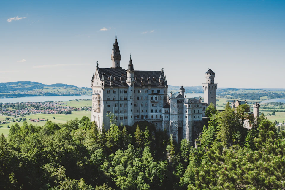 The image depicts a majestic castle perched atop a hill, surrounded by lush greenery and trees. The castle features multiple towers and spires that reach towards the clear blue sky. The perspective of the photo is from below, looking up at the castle, which enhances its grandeur and prominence in the landscape.