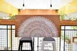 A large decorative wooden panel with intricate white mandala patterns rests on two stools. The backdrop consists of a modern interior with bright yellow walls, black grid panels, and several potted plants. Two hanging light bulbs can be seen above the panel.
