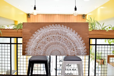 A large decorative wooden panel with intricate white mandala patterns rests on two stools. The backdrop consists of a modern interior with bright yellow walls, black grid panels, and several potted plants. Two hanging light bulbs can be seen above the panel.