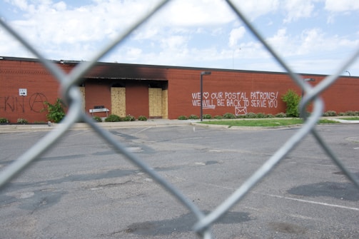 A chain-link fence in the foreground frames a view of a large brick building with boarded-up windows and a spray-painted message. The building is surrounded by an empty parking lot and sparse greenery. The message on the brick wall reads, 'WE ♥ OUR POSTAL PATRONS! WE WILL BE BACK TO SERVE YOU'.