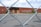 A chain-link fence in the foreground frames a view of a large brick building with boarded-up windows and a spray-painted message. The building is surrounded by an empty parking lot and sparse greenery. The message on the brick wall reads, 'WE ♥ OUR POSTAL PATRONS! WE WILL BE BACK TO SERVE YOU'.