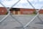 A chain-link fence in the foreground frames a view of a large brick building with boarded-up windows and a spray-painted message. The building is surrounded by an empty parking lot and sparse greenery. The message on the brick wall reads, 'WE ♥ OUR POSTAL PATRONS! WE WILL BE BACK TO SERVE YOU'.
