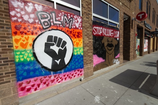 Murals are painted on the walls of a brick building, featuring a Black Lives Matter theme. On the left, a rainbow background is filled with colorful hearts and the letters 'BLM' above a large black fist. On the right, a portrait of a person's face partially covered by the words 'Stop Killing Us!' in bold red letters.
