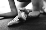 A close-up of a hand in a yoga pose, with fingers forming a mudra gesture. The focus is on the fingers, which rest on a textured mat. The background is blurred, suggesting an indoor setting.