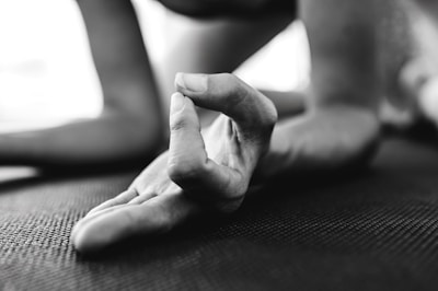 A close-up of a hand in a yoga pose, with fingers forming a mudra gesture. The focus is on the fingers, which rest on a textured mat. The background is blurred, suggesting an indoor setting.