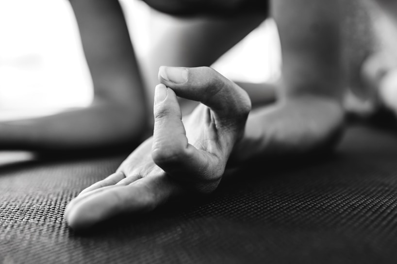 A close-up of a hand in a yoga pose, with fingers forming a mudra gesture. The focus is on the fingers, which rest on a textured mat. The background is blurred, suggesting an indoor setting.