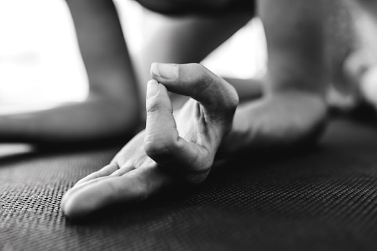 A close-up of a hand in a yoga pose, with fingers forming a mudra gesture. The focus is on the fingers, which rest on a textured mat. The background is blurred, suggesting an indoor setting.